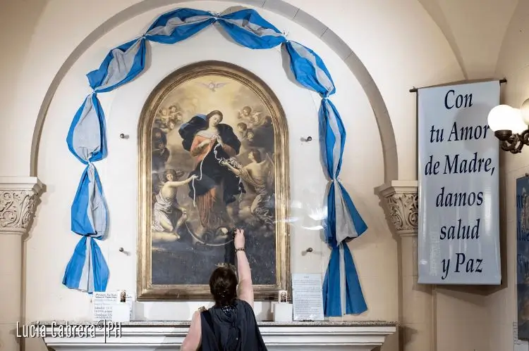 La imagen muestra una pintura de la Virgen María Desatanudos, ubicada en la Parroquia San José del Talar en Buenos Aires, un santuario popular. El texto visible en un cartel lateral dice: "Con tu Amor de Madre damos salud y Paz". 