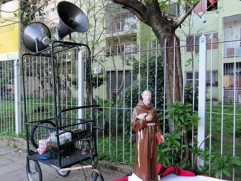 La imagen muestra una estatua de San Francisco de Asís colocada junto a un carro con altavoces en un entorno urbano, de villa soldati