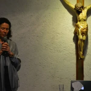Esta fotografía muestra a un laico consagrado hablando frente a un micrófono junto a un crucifijo, con una estatua dorada de Jesucristo montada en una cruz de madera en el fondo. La escena parece ser parte de un evento religioso o espiritual, con una laptop y otros objetos pequeños visibles sobre la mesa.