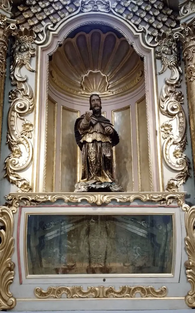 Esta imagen muestra una figura de santo dentro de un retablo barroco en la Parroquia de San Ignacio de Loyola en Buenos Aires.La escultura está situada en una hornacina con forma de venera (concha) gallonada.La figura tallada corresponde específicamente a San Juan Nepomuceno, considerado el patrón de los confesores.